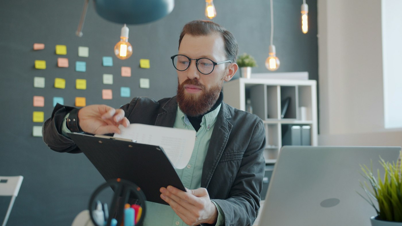 A compliance lead reviews documents at a desk in front of a planning wall covered in tracking notes.