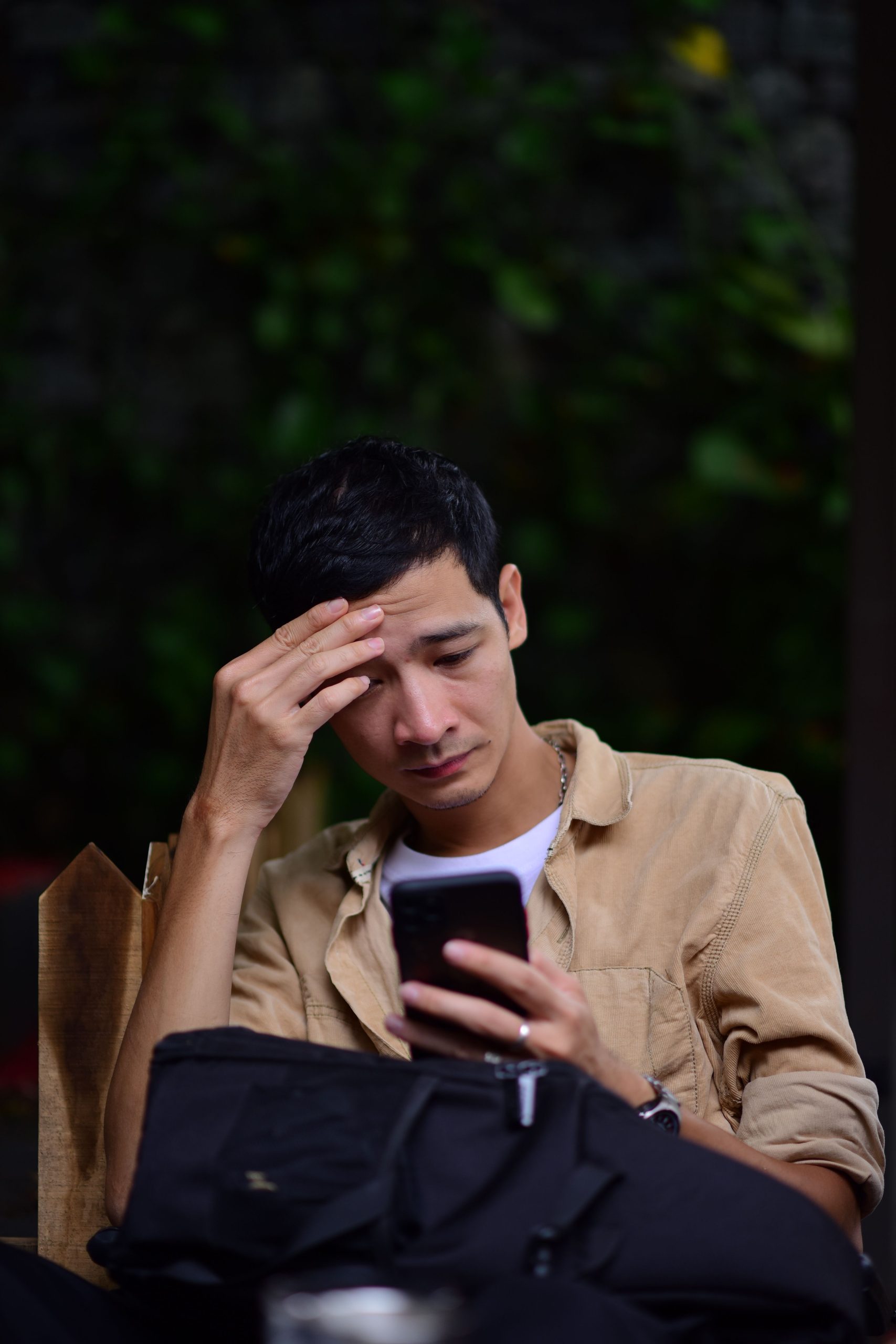 A young man sitting outdoors reading a text message on his phone with a worried expression